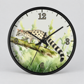 A round wall clock with a black frame and a printed watercolor-style illustration of a spotted cat walking along a branch in green grass. The clock face has black numeral markers and black hands, shown against a light gray background.