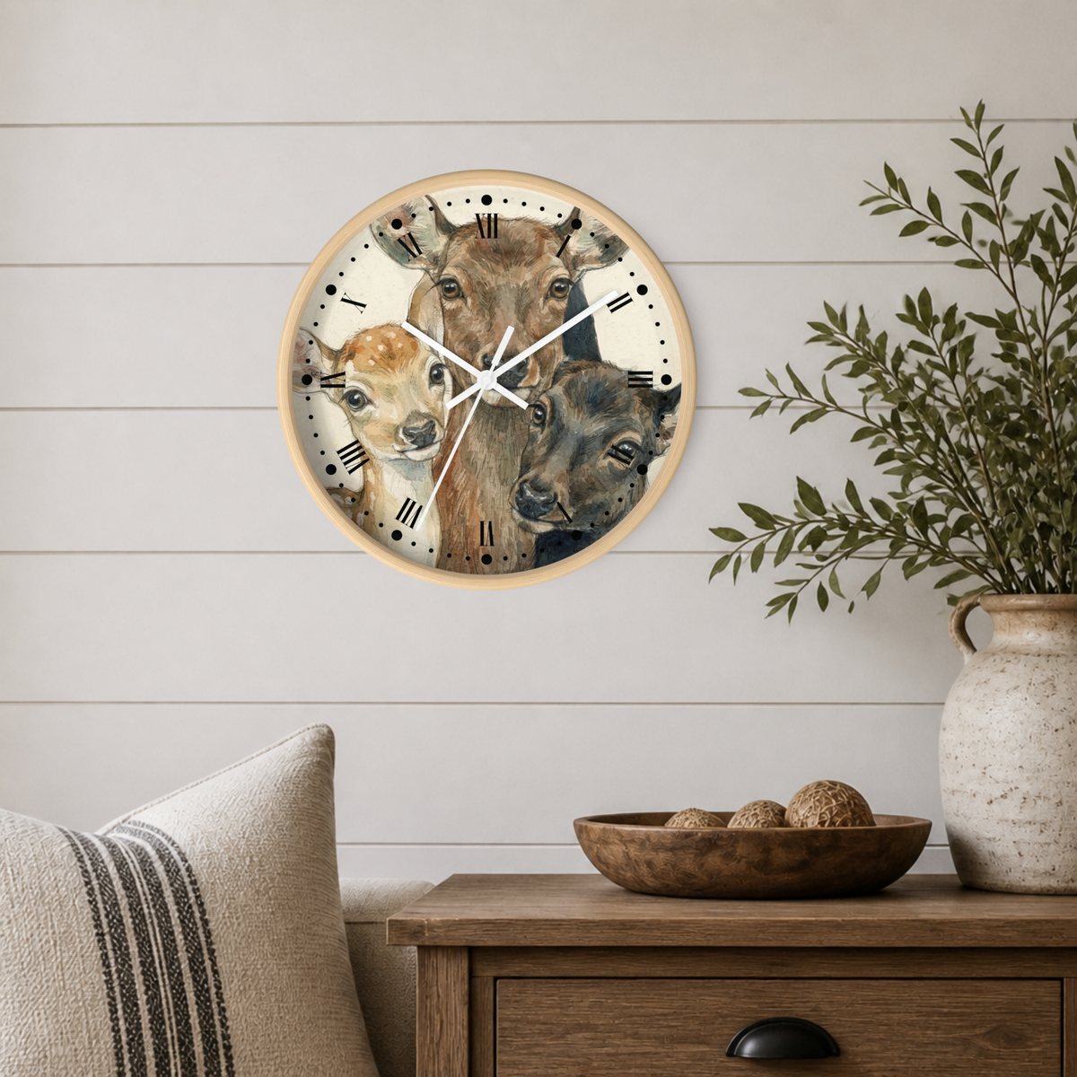 A round wall clock with a light wood frame and a printed illustration of three deer on the face, mounted on a white shiplap wall above a wooden side table with a vase and bowl.