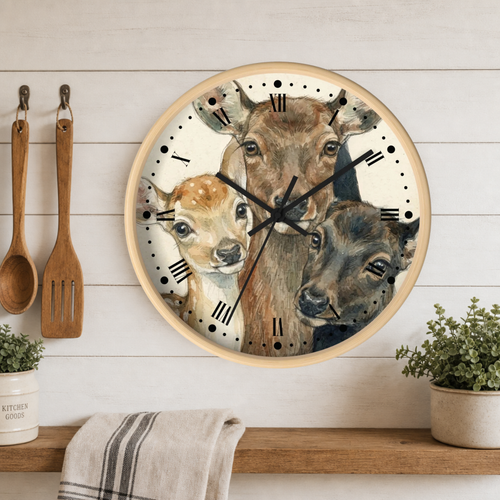 Round wall clock with a watercolor-style illustration of three deer on the face, mounted on a white wood plank wall above a wooden shelf with plants and a towel. A wooden spoon and spatula hang on the left.