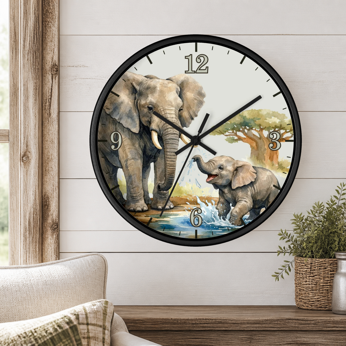 A round wall clock with a black frame and a printed safari design featuring an adult and baby elephant in a watering hole, mounted on a white wood-paneled wall above a wooden console table with a small potted plant.