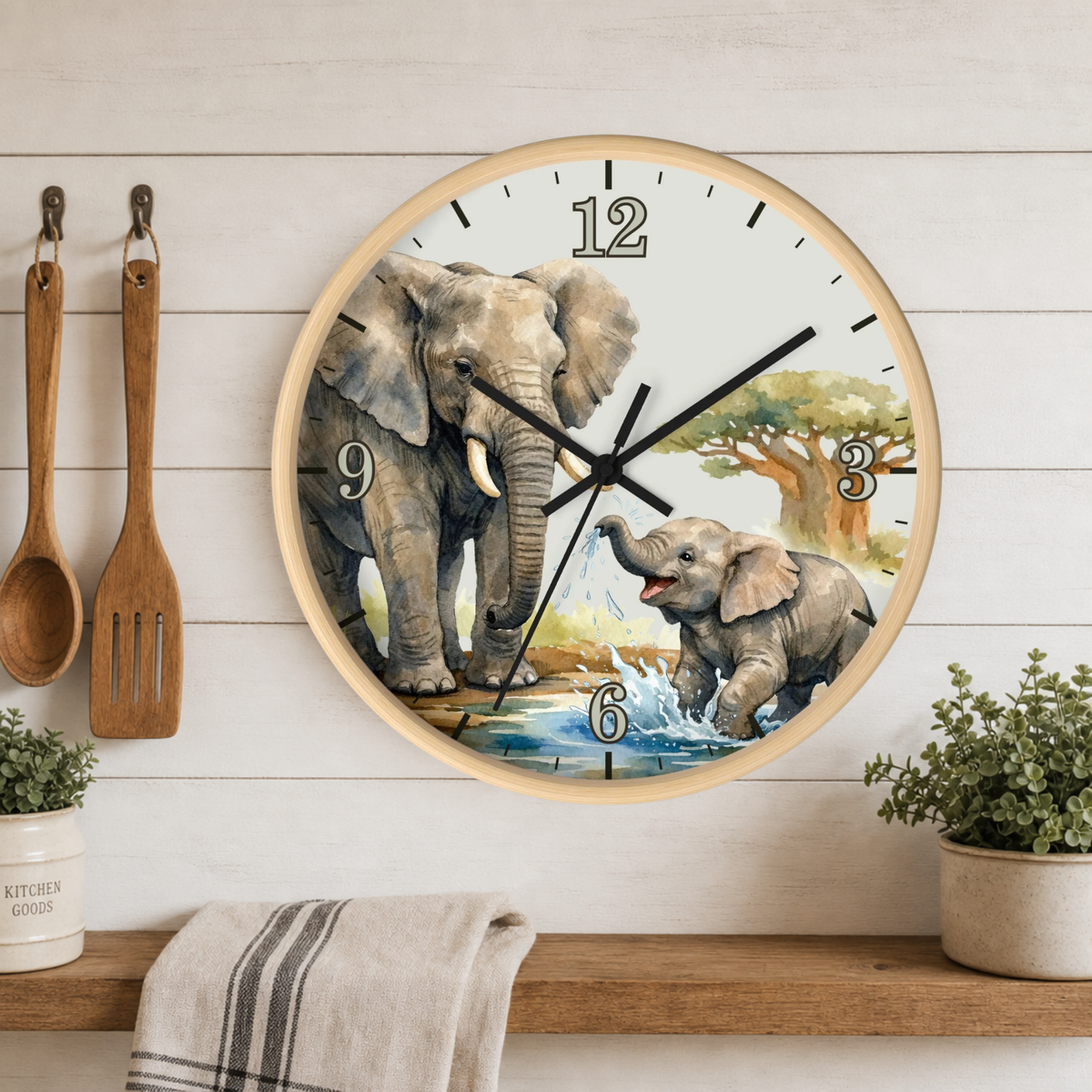 A round wall clock with a light wood frame and a watercolor-style elephant illustration on the face, hanging on a white wood-paneled wall above a shelf with plants and kitchen utensils.