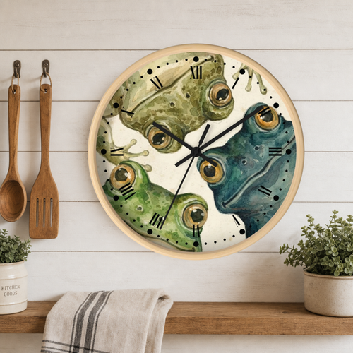 Round wall clock with a watercolor design of two frogs facing each other, one green and one blue, on a white wood-paneled kitchen wall above a wooden shelf with a towel and small plants.