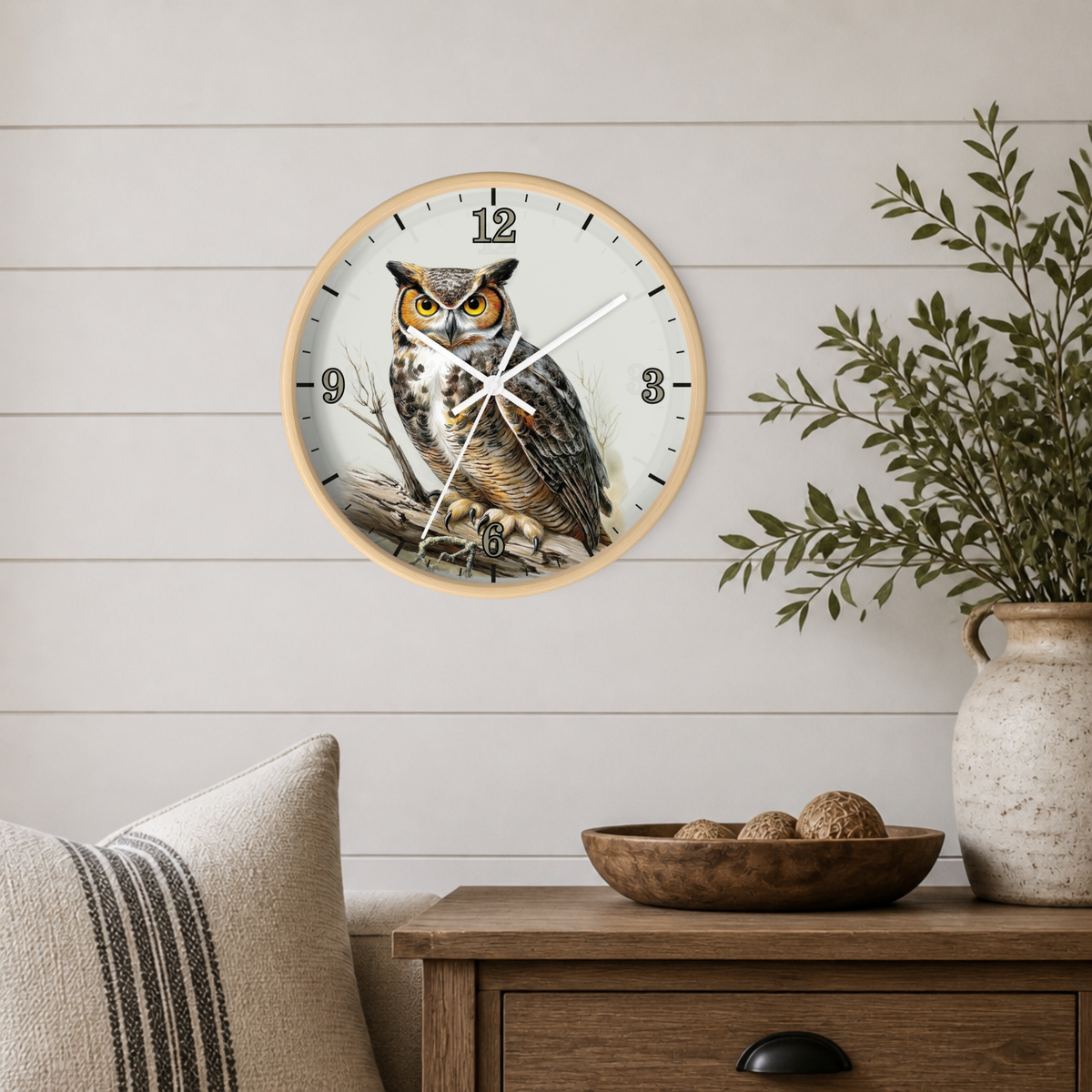 A round wall clock featuring a detailed illustration of a great horned owl on the face, mounted on a white shiplap wall above a wooden side table with a vase and bowl.