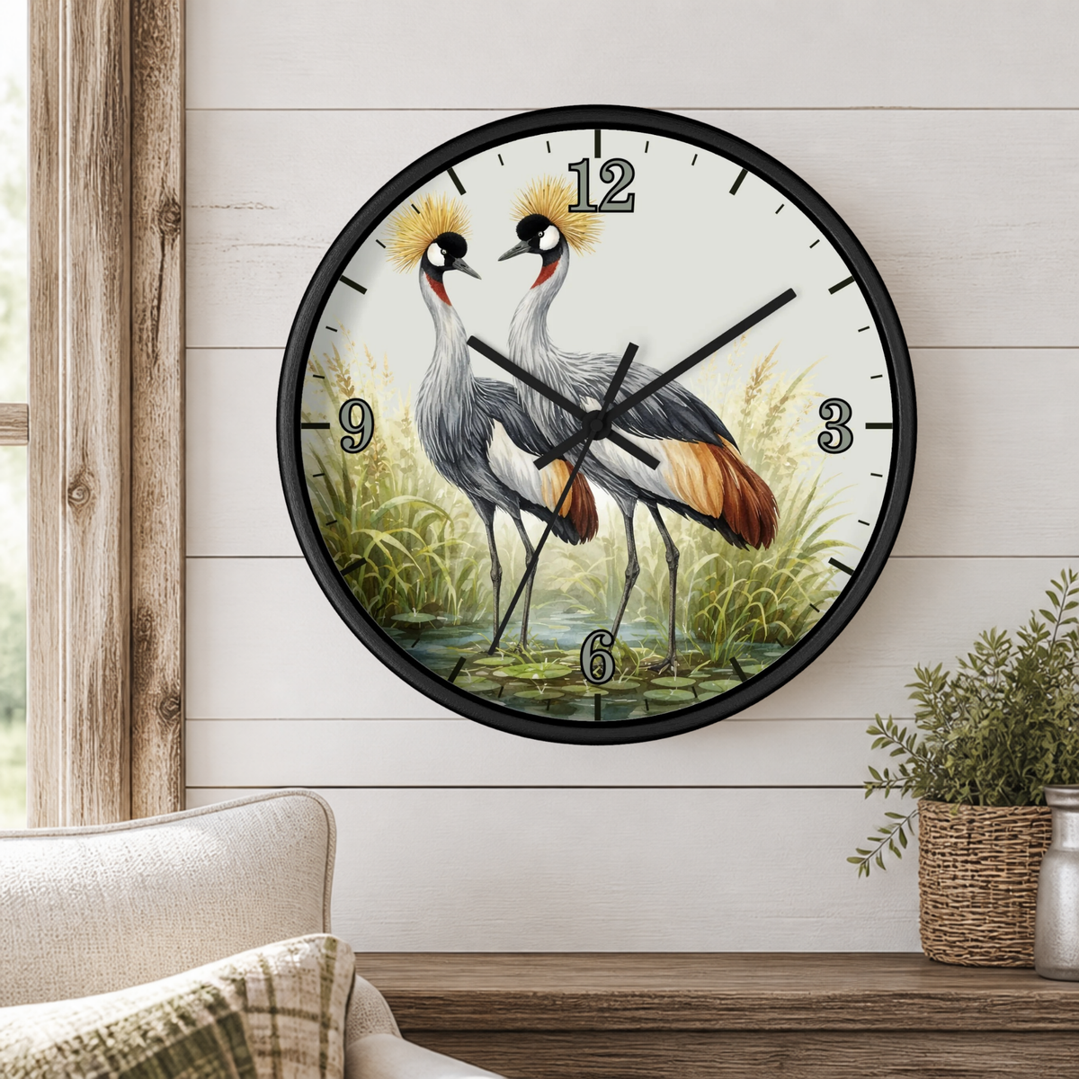 Round wall clock with a black frame and a printed illustration of two crowned cranes on the face, mounted on a white shiplap wall above a wooden console table with a plant and part of a sofa visible.