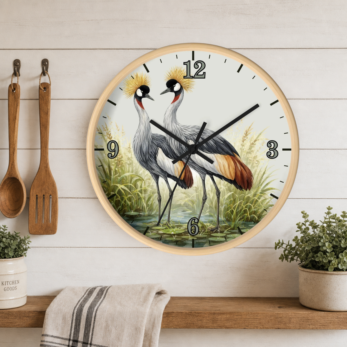 Round wall clock with a light wood frame featuring a detailed illustration of two crowned cranes standing in water. The clock is mounted on a white wood-paneled wall above a wooden shelf with kitchen utensils, a towel, and small potted plants.