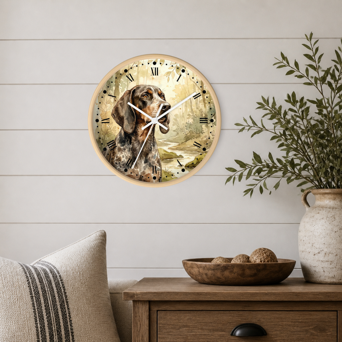 A round wall clock with a light wood frame hangs on a white shiplap wall above a wooden side table. The clock face features a vintage-style illustration of a hunting dog in warm beige, brown, and cream tones with black Roman numerals.