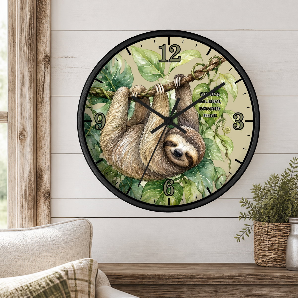A round wall clock with a black frame and a sloth illustration on the face, mounted on a white wood-paneled wall above a wooden table with a small potted plant.