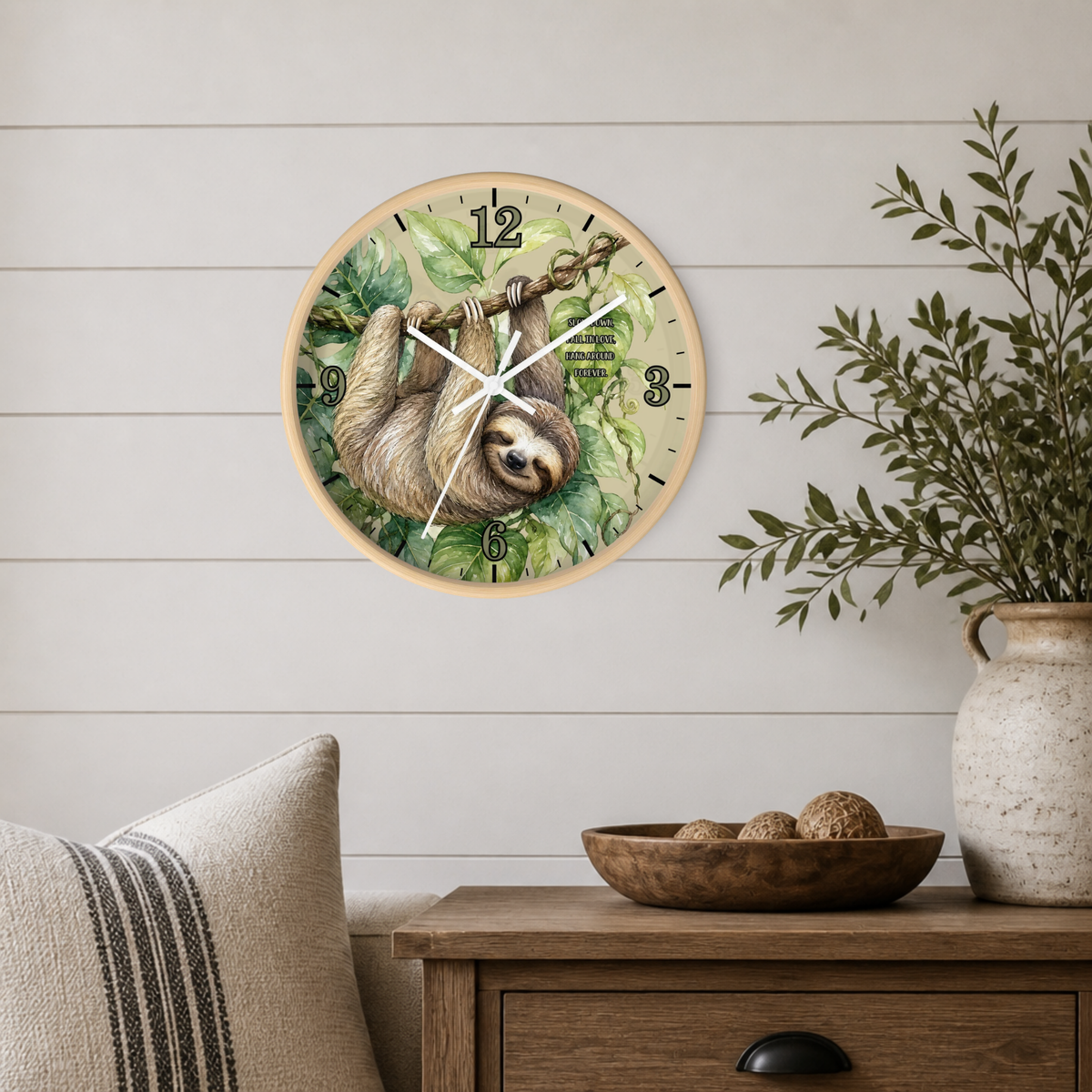 A round wall clock featuring a sloth illustration on a leafy green background, mounted on a white shiplap wall above a wooden side table with a vase and bowl.