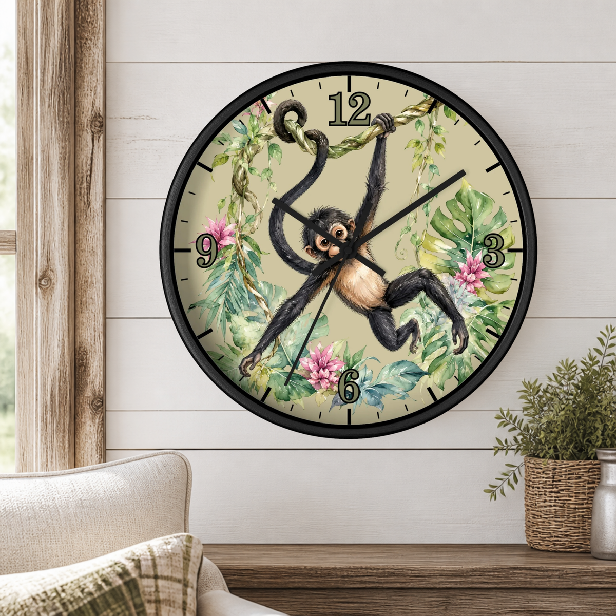 A round wall clock with a black frame and a whimsical illustration of a hanging monkey among green leaves and pink flowers on a cream background, mounted on a white wood-paneled wall above a wooden table with a plant and part of a sofa visible.