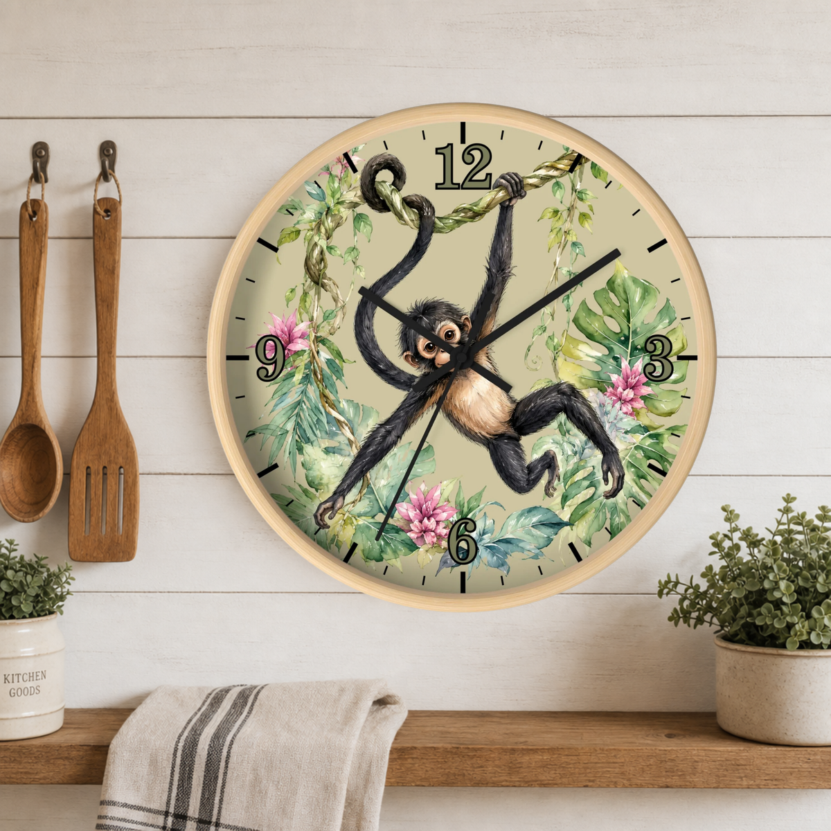 A round wall clock with a tropical jungle design featuring a hanging chimpanzee, green leaves, pink flowers, and black numerals, mounted on a white wood-paneled wall above a shelf with kitchen utensils and plants.