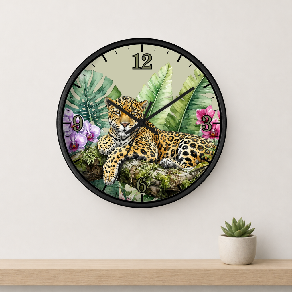 A round wall clock with a black frame and a tropical jungle design featuring a leopard, palm leaves, and pink flowers. The clock is mounted on a white wall above a light wood shelf with a small potted succulent.