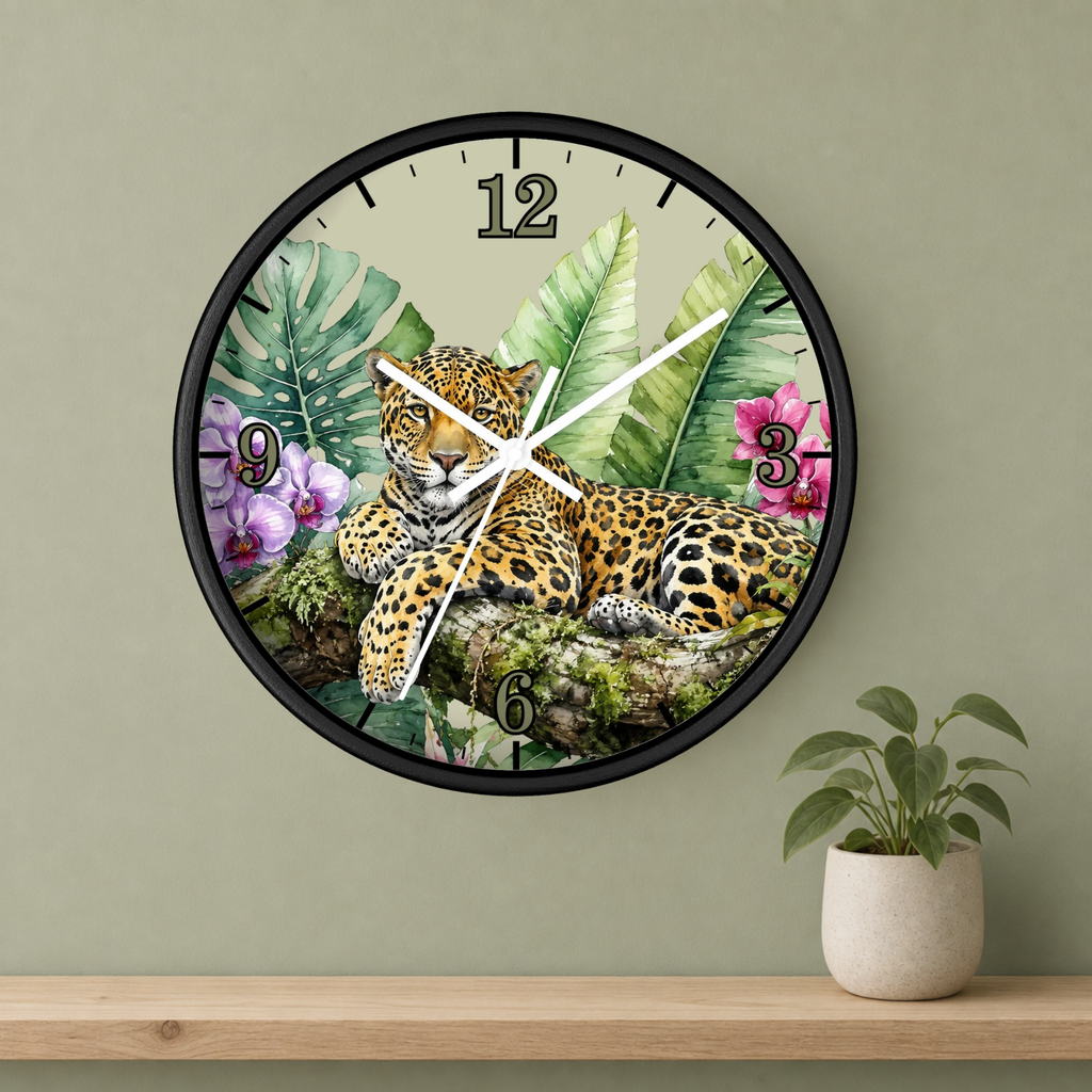 A round wall clock with a black frame and a tropical jungle print featuring a leopard, palm leaves, and pink flowers. The clock is mounted on a sage green wall above a wooden shelf with a small potted plant.