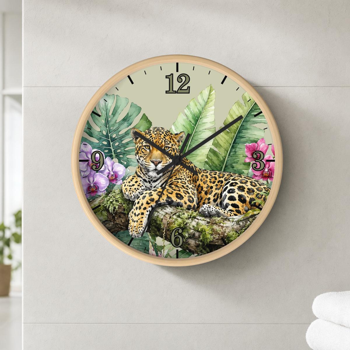 A round wall clock with a light wood frame, featuring a detailed illustration of a leopard resting among tropical green leaves and pink flowers. The clock face has black Arabic numerals and black hands, and it is mounted on a light gray tiled wall.