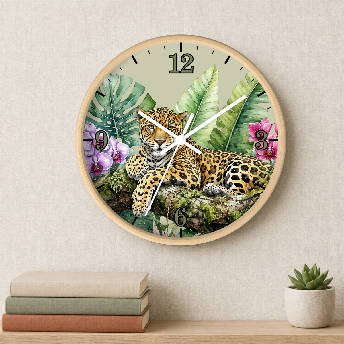 A round wall clock with a light wood frame hangs on a beige wall above a shelf with stacked books and a small potted succulent. The clock face features a detailed illustration of a leopard lying among tropical green leaves and pink flowers.