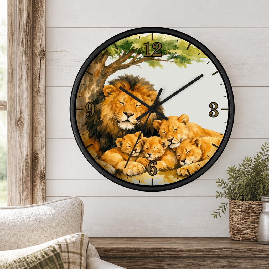 A round wall clock with a black frame and a printed lion family illustration on the face, mounted on a white wood-paneled wall above a wooden surface with a small plant and part of a chair visible.