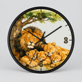 A round wall clock with a black frame and a printed illustration of a lion family under a tree. The clock face has a white background with black hour markers and black hands.