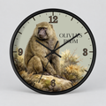A round wall clock with a black frame and a printed illustration of a baboon sitting on rocks, with the text 'OLIVIA'S ROOM' on the clock face. The clock is photographed against a neutral light gray background.