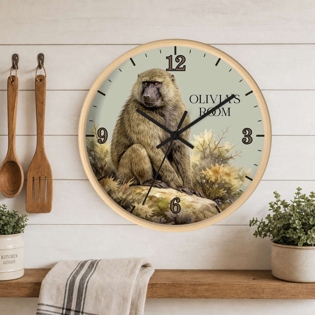 A round wall clock with a light wood frame hangs on a white wood-paneled wall above a wooden shelf. The clock face features a detailed illustration of a brown baboon sitting on rocks with the text 'OLIVIA'S ROOM' and black Arabic numerals.