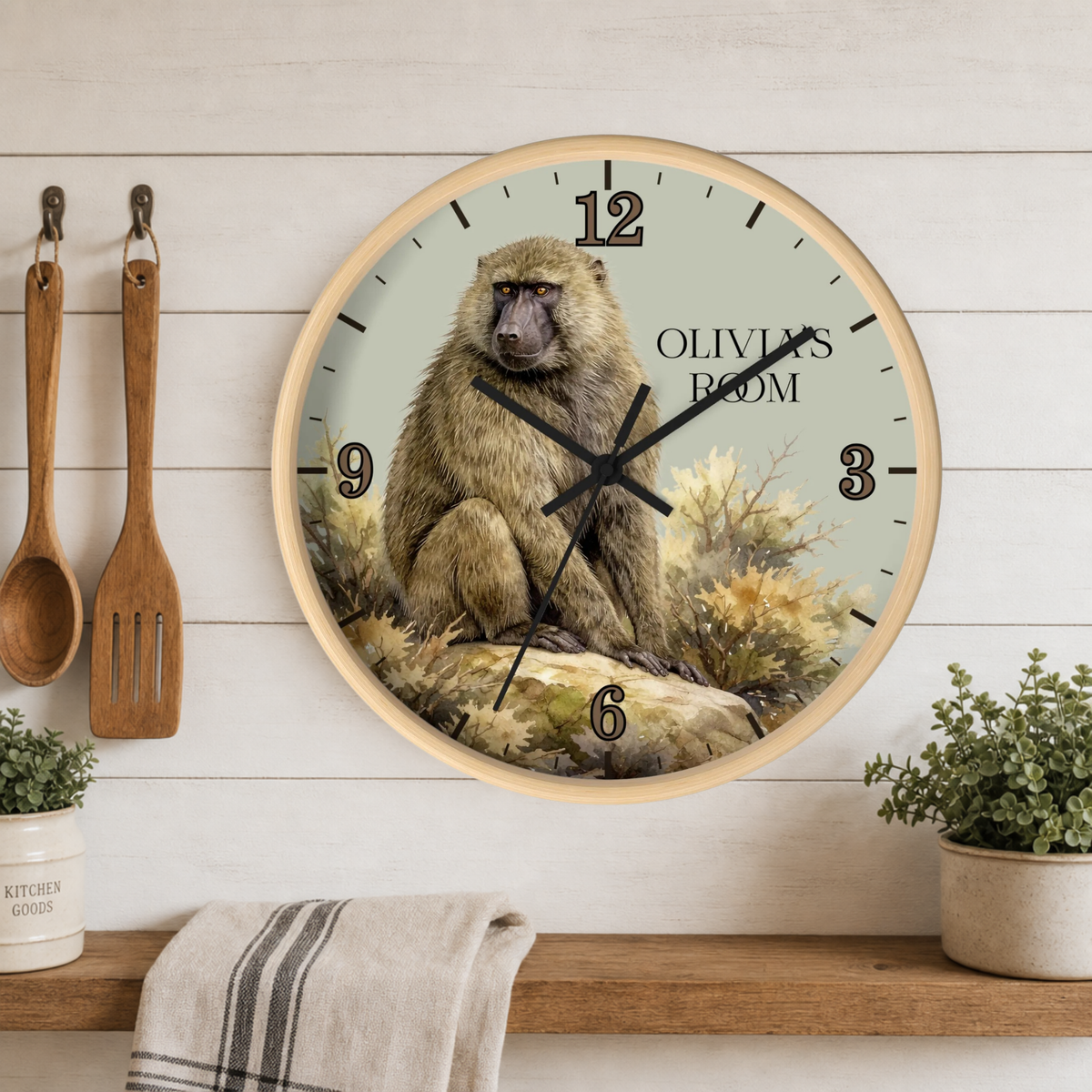A round wall clock with a light wood frame hangs on a white wood-paneled wall above a wooden shelf. The clock face features a detailed illustration of a brown baboon sitting on rocks with the text 'OLIVIA'S ROOM' and black Arabic numerals.