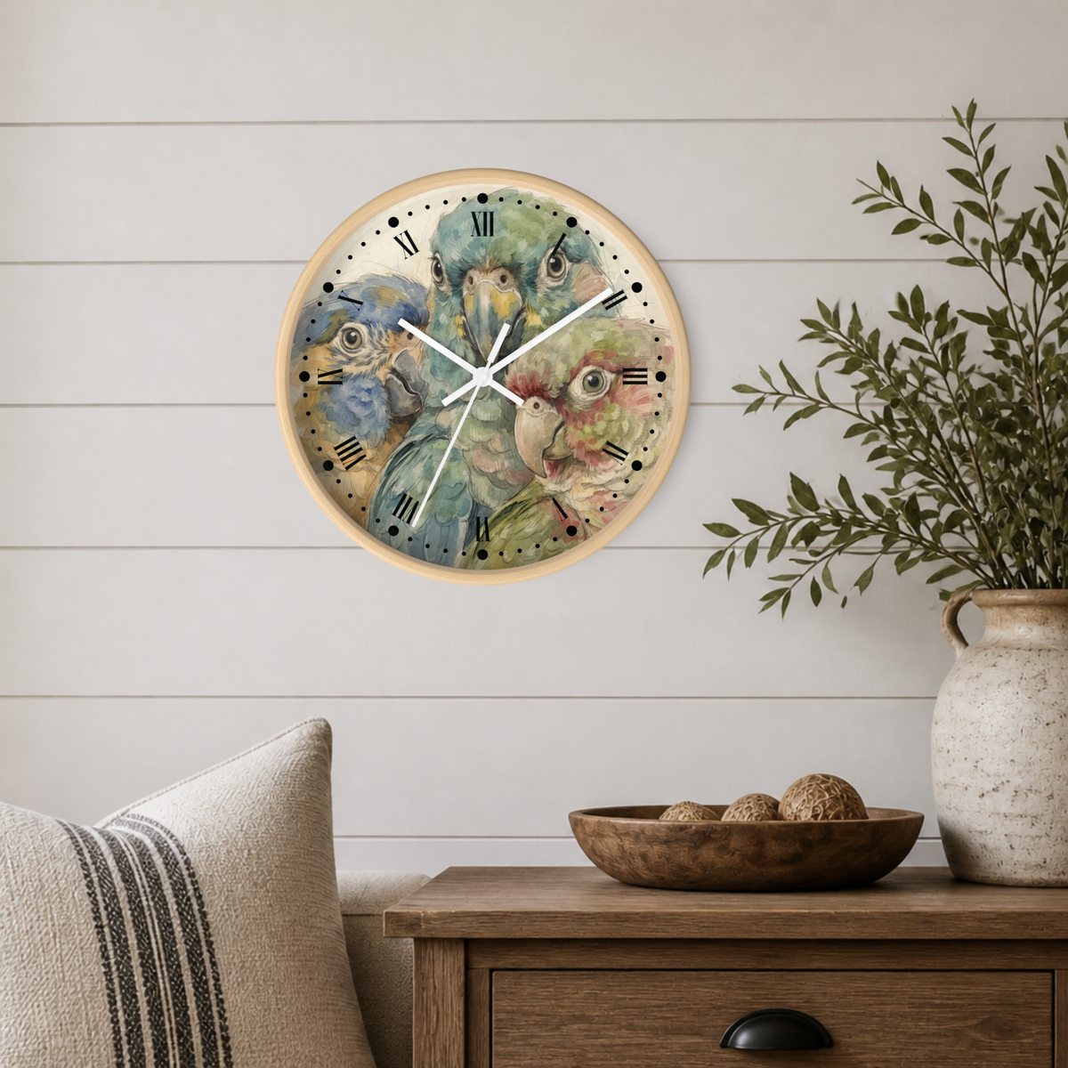 A round wall clock with a light wood frame and a colorful watercolor design of parrots on the face, mounted on a white shiplap wall above a wooden side table with a plant and decorative items.