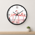 A round wall clock with a black frame and a white face featuring three pink flamingos standing in shallow water. The clock is mounted on a light beige wall above a wooden shelf with a small potted succulent.