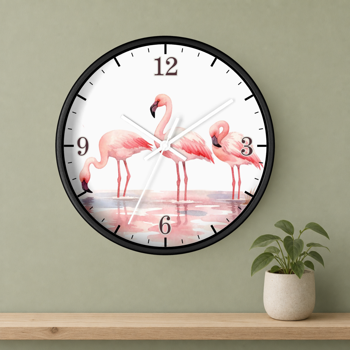 A round wall clock with a black frame and a white face featuring three pink flamingos standing in shallow water. The clock is mounted on a sage green wall above a light wood shelf with a small potted plant.