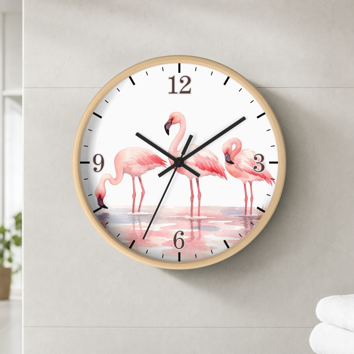A round wall clock with a light wood frame and a white face printed with pink flamingos standing in shallow water. The clock is mounted on a light gray wall.