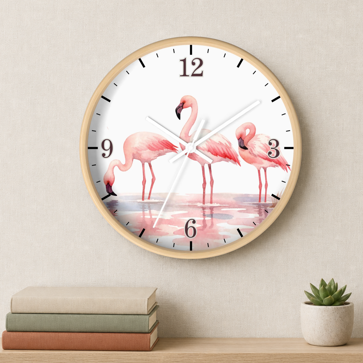 A round wall clock with a light wood frame and a white face featuring three pink flamingos standing in shallow water. The clock is mounted on a beige wall above a wooden shelf holding stacked books and a small potted succulent.