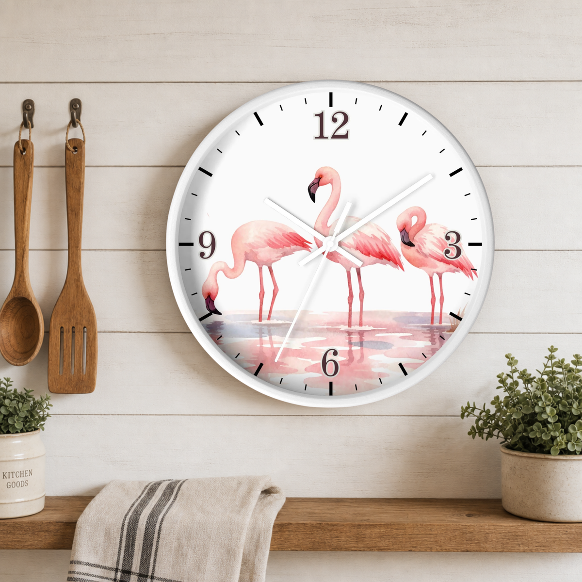 A round wall clock with a white face and black numerals, featuring a watercolor-style illustration of three pink flamingos standing in shallow water. The clock is mounted on a light wood-paneled wall above a shelf with kitchen utensils, plants, and a towel.