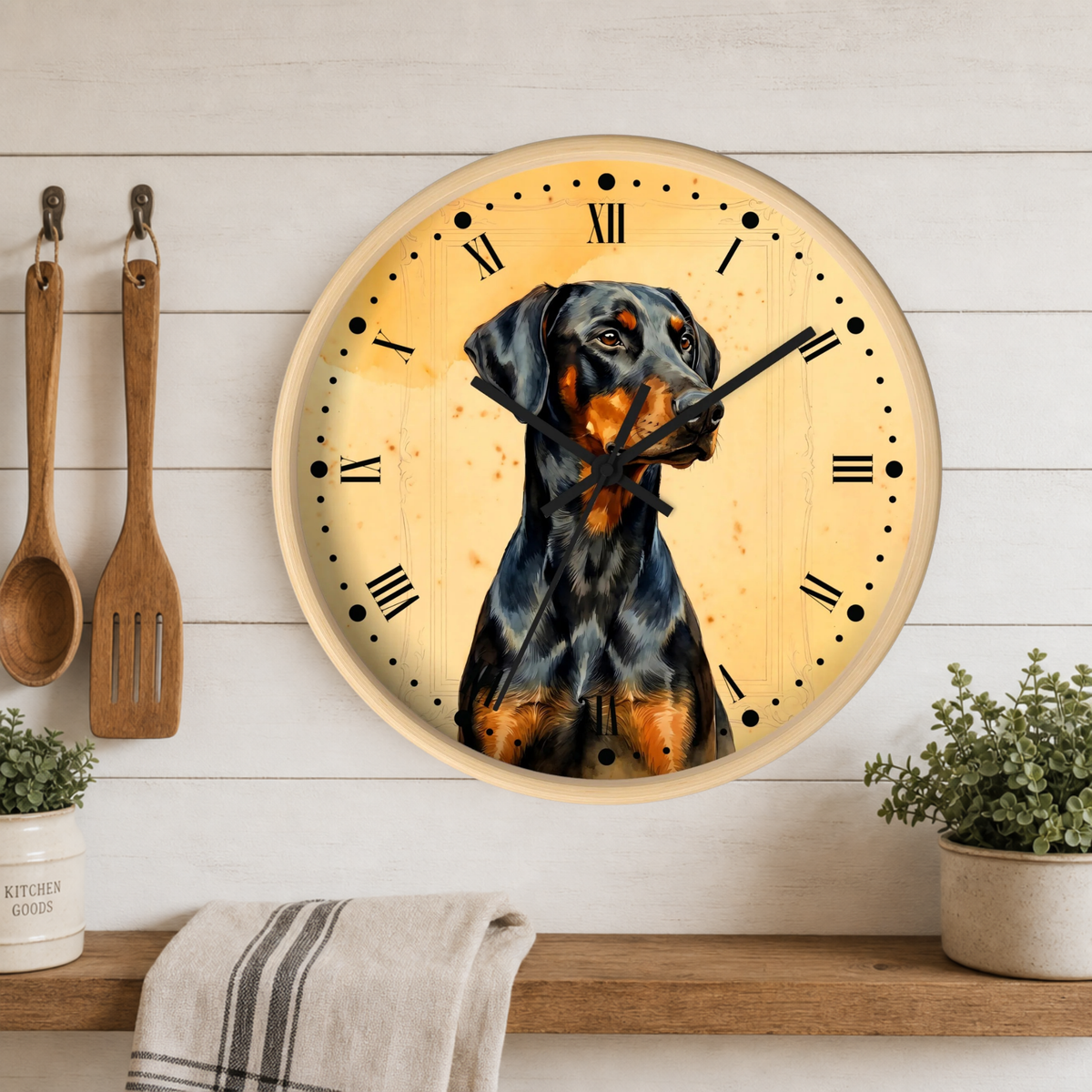 A round wall clock with a light wood frame and a cream-colored face featuring a painted  dog. The clock is mounted on a white wood-paneled wall above a wooden shelf with kitchen utensils, a towel, and small potted plants.