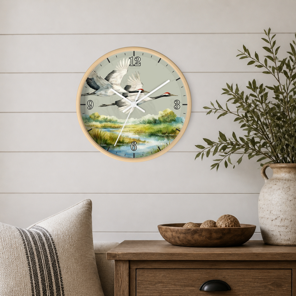 A round wall clock with a natural wood frame and a scenic print of flying cranes over a marsh landscape, mounted on a white shiplap wall above a wooden side table with a plant and decorative bowl.
