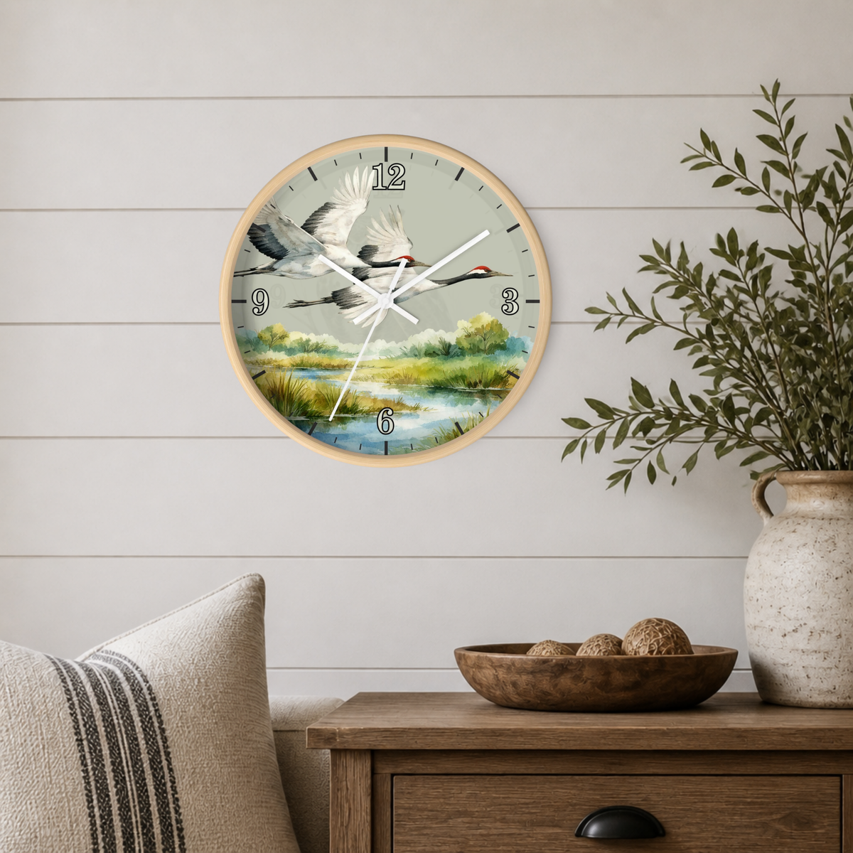 A round wall clock with a natural wood frame and a scenic print of flying cranes over a marsh landscape, mounted on a white shiplap wall above a wooden side table with a plant and decorative bowl.