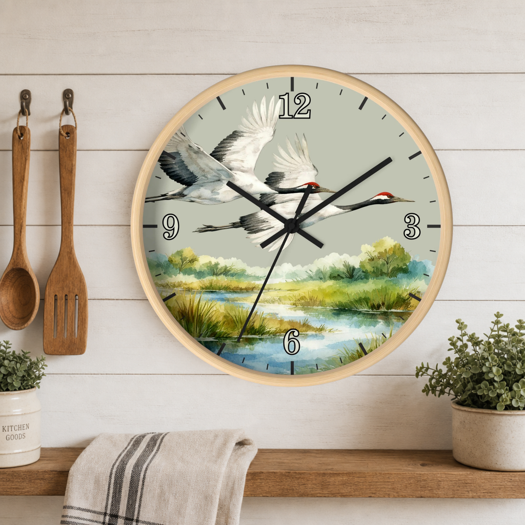 Round wall clock with a light wood frame featuring a painted scene of white cranes flying over a marsh landscape. The clock is mounted on a white wood-paneled wall above a wooden shelf with kitchen utensils, a towel, and potted plants.