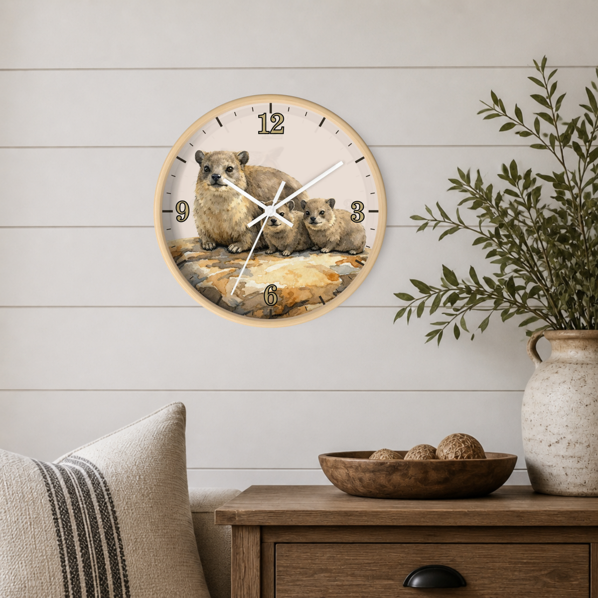 A round wall clock with a natural wood frame hangs on a white shiplap wall above a wooden side table. The clock face features a watercolor illustration of rock hyrax and two pups on a rock, with black Arabic numerals and white hands.