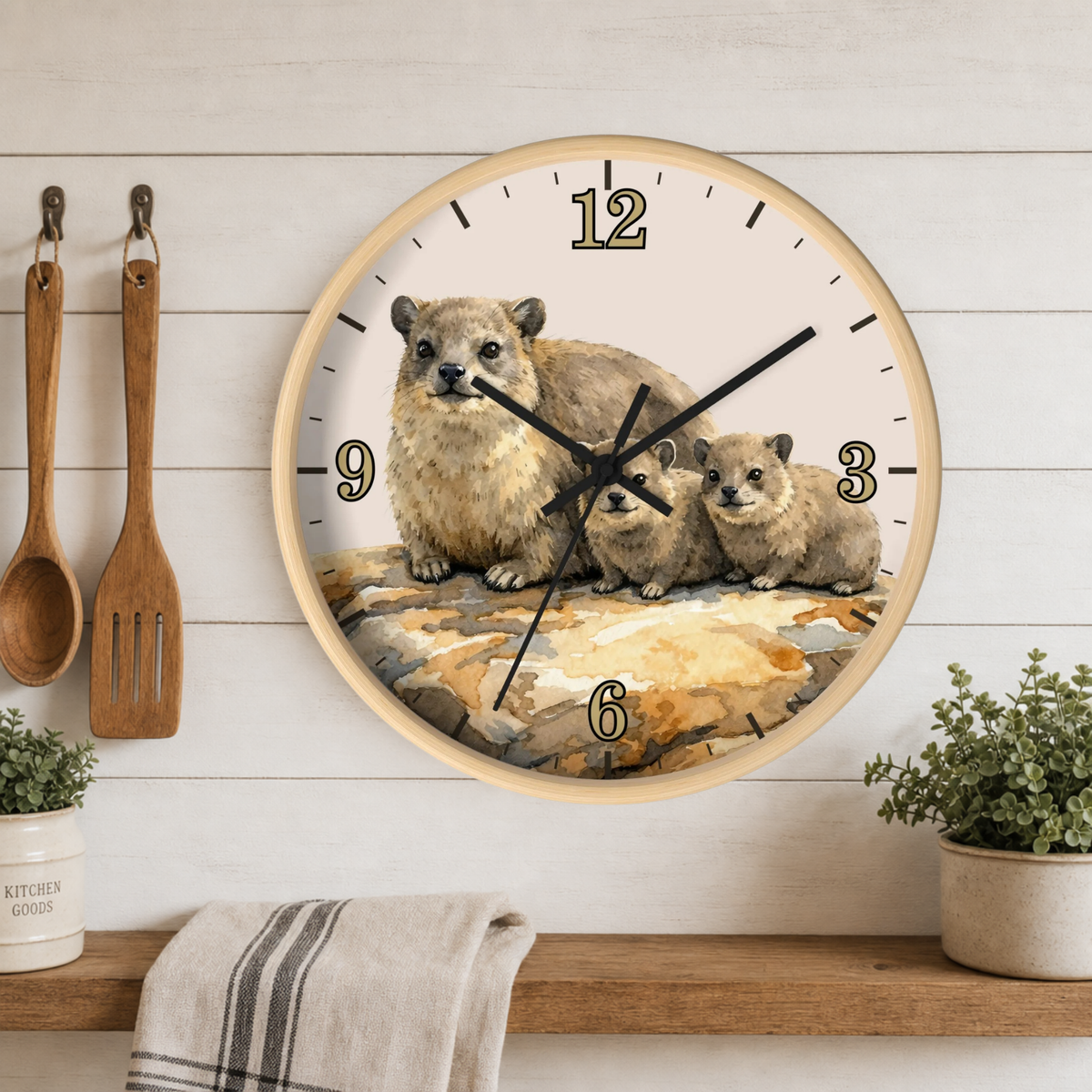A round wall clock with a light wood frame and a printed illustration of a mother rock hyrax with two pups on the clock face, mounted on a white wood-paneled wall above a shelf with kitchen utensils and plants. 