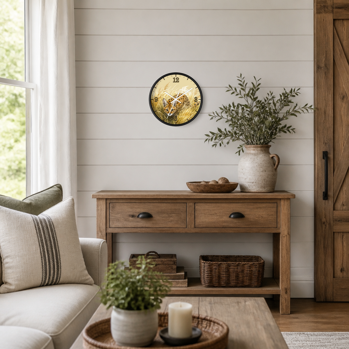 A round wall clock featuring a watercolor illustration of a spotted serval cat in tall grass, styled in a cozy, light-colored living room above a wooden console table.