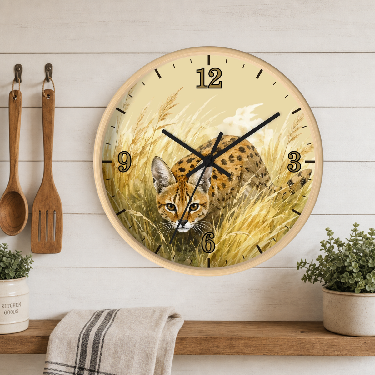 Round wall clock with a natural wood frame, featuring a printed design of a spotted big cat in tall golden grass. The clock is mounted on a white wood paneled wall above a wooden shelf with kitchen utensils, a towel, and small potted plants.