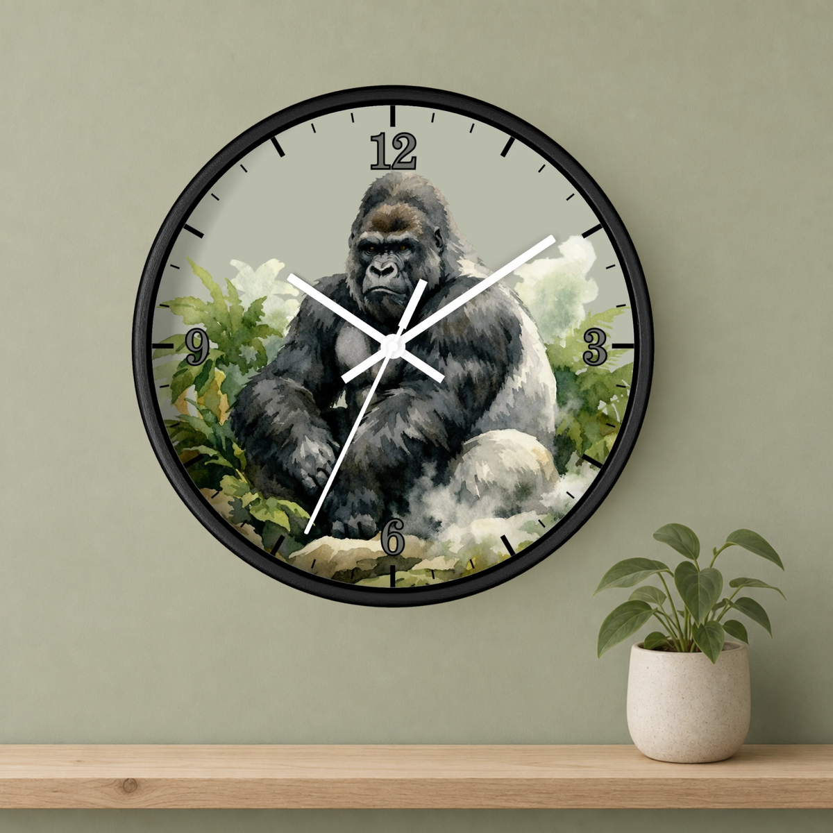 Round wall clock with a black frame and a printed gorilla illustration on the face, mounted on a sage green wall above a wooden shelf with a small potted plant.