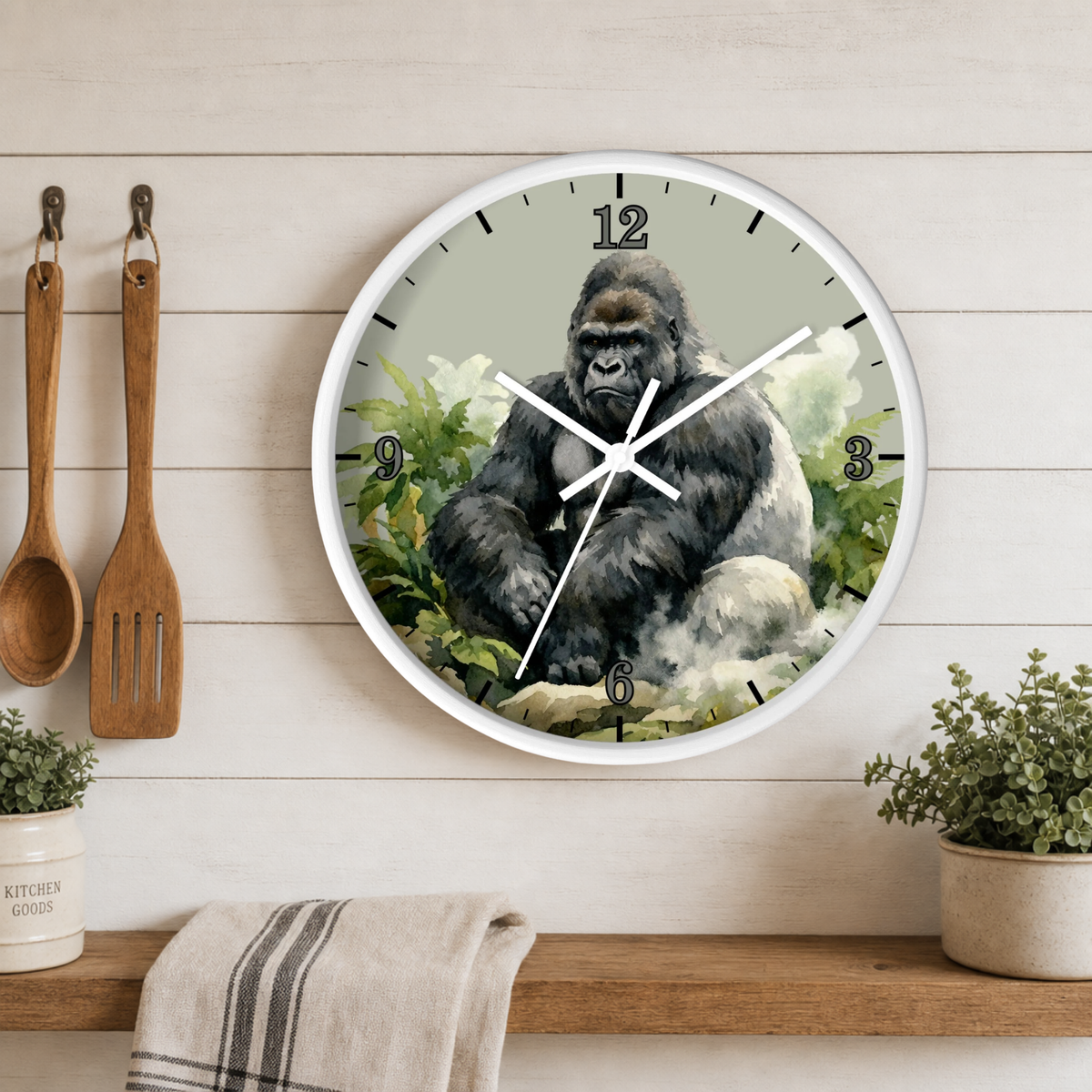 A round wall clock with a white frame hangs on a light wood plank wall above a shelf. The clock face features a detailed illustration of a gorilla seated among green foliage.