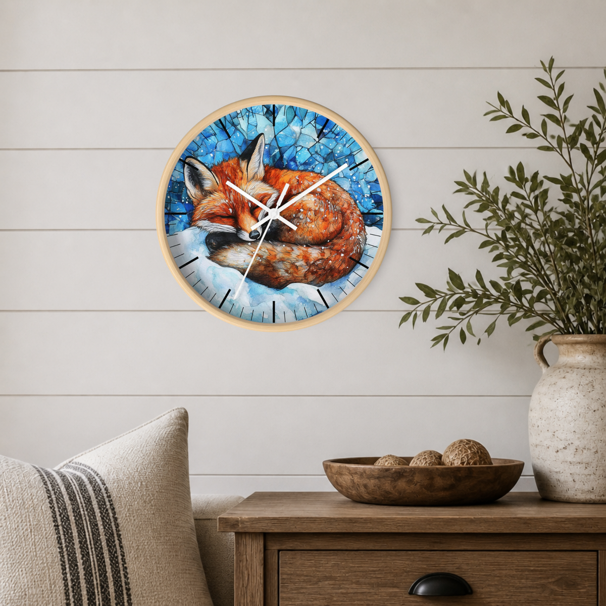 A round wall clock with a light wood frame hangs on a white shiplap wall above a wooden side table. The clock face features a colorful illustration of a sleeping fox in a blue mosaic-style background.
