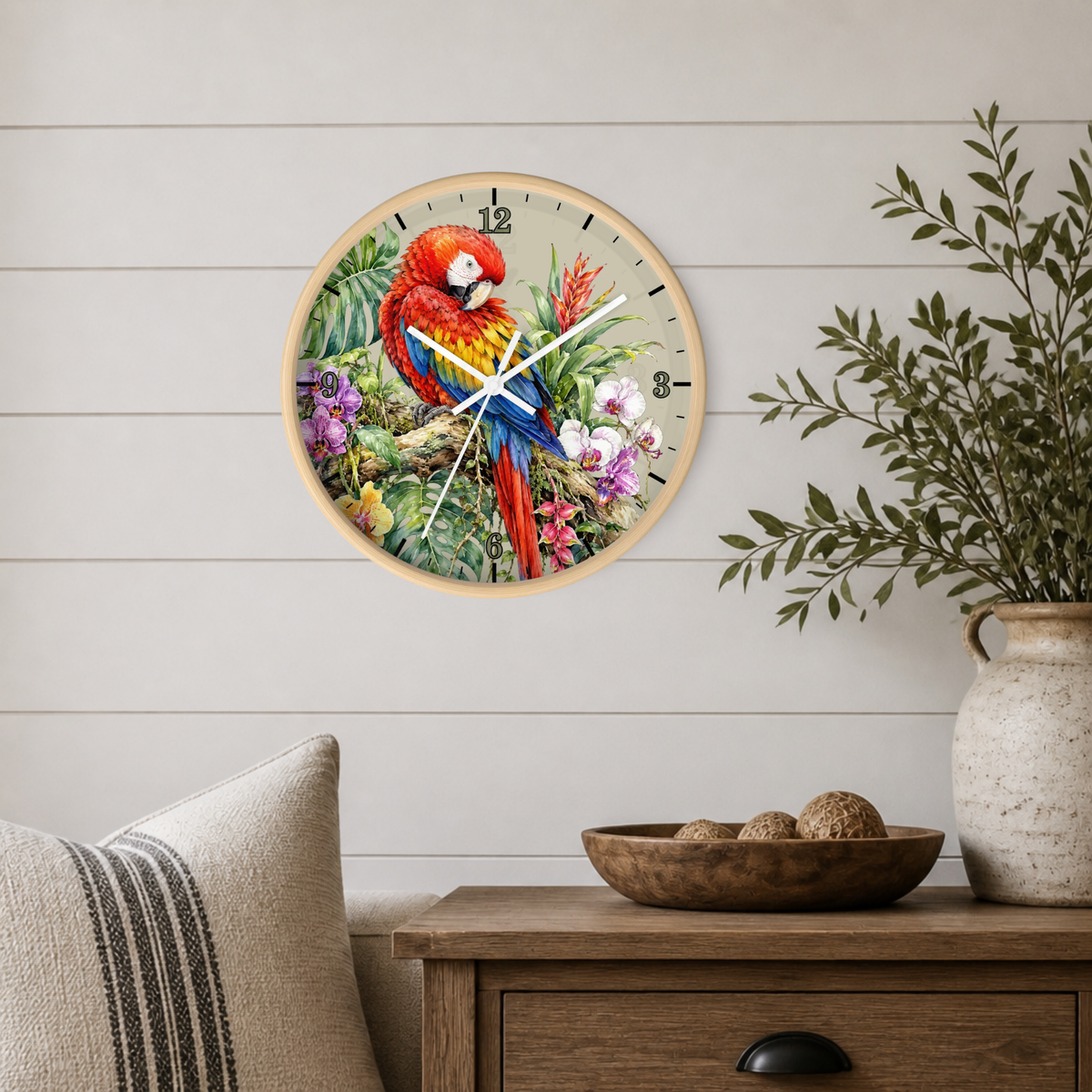 A round wall clock with a light wood frame hangs on a white shiplap wall above a wooden side table. The clock face features a colorful illustration of a red and blue parrot perched among tropical flowers.