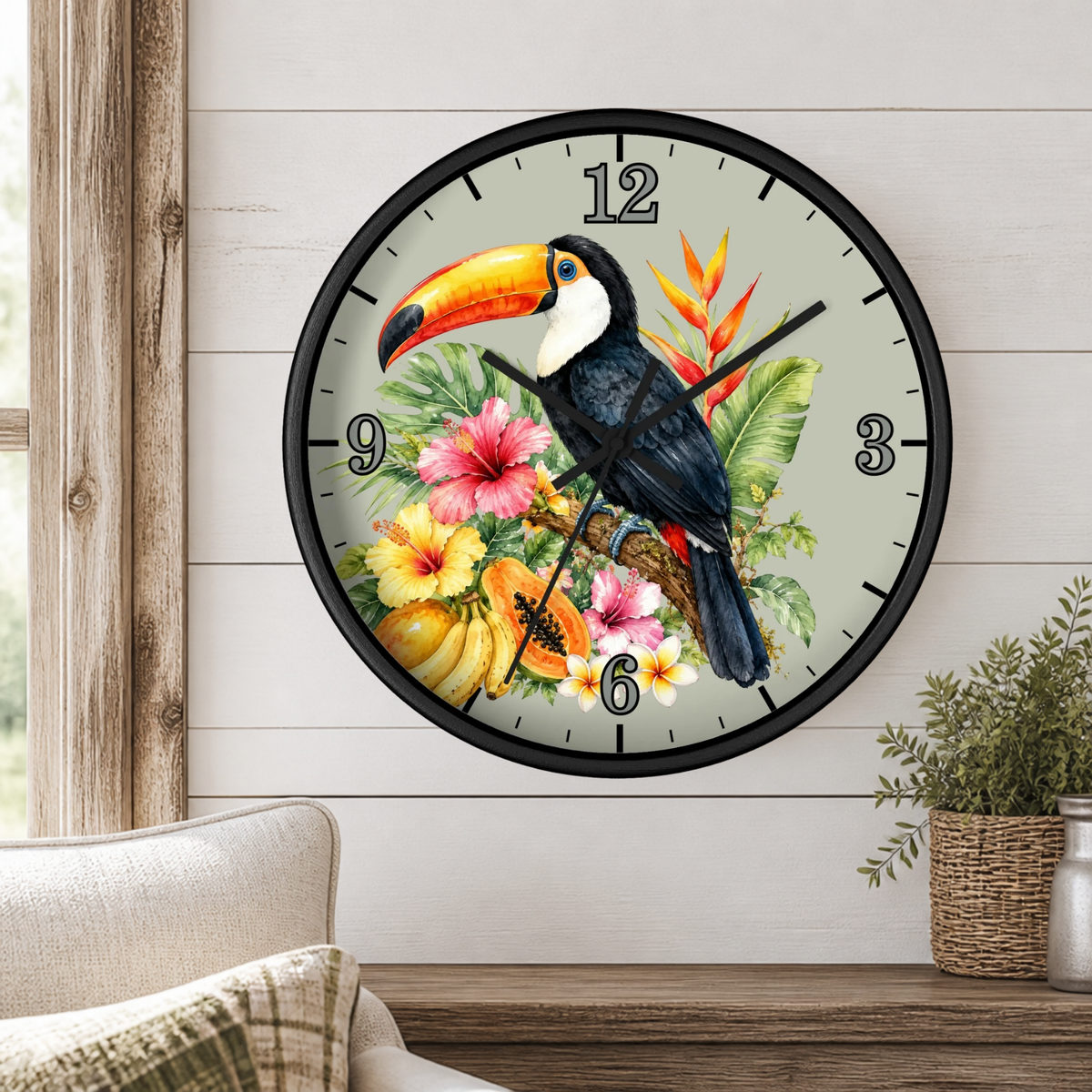 A round wall clock with a black frame and a tropical toucan and floral design on the face, mounted on a white wood-paneled wall above a wooden shelf with a small potted plant.