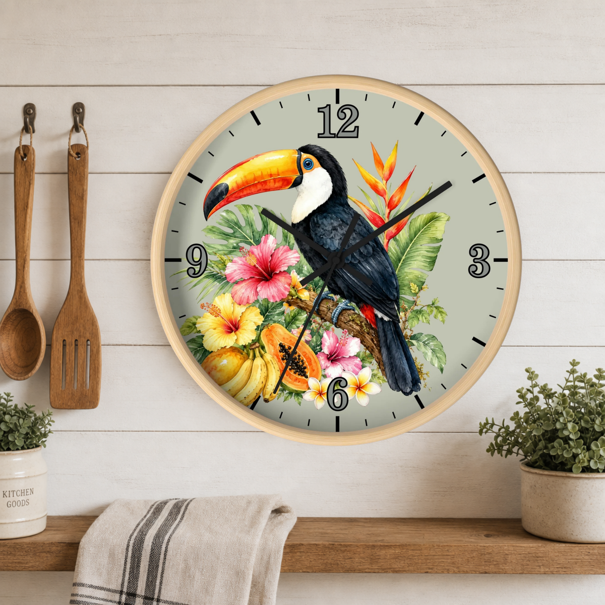 A round wall clock with a light wood frame hangs on a white wood plank wall above a shelf. The clock face features a colorful illustration of a toucan, hibiscus flowers, and tropical leaves.