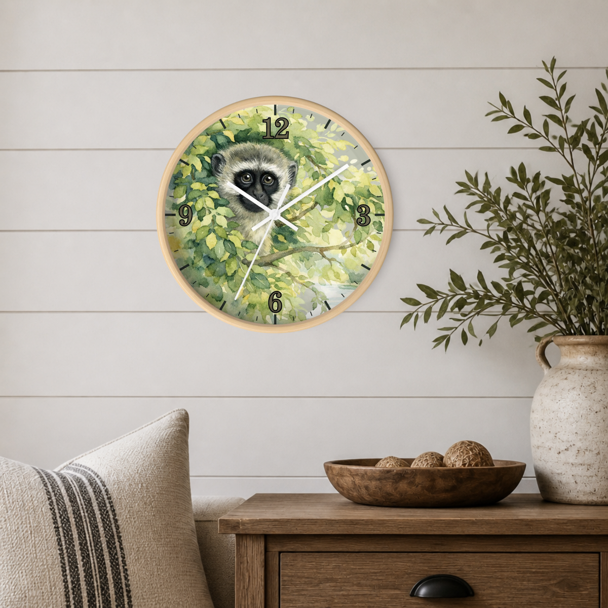 Round wall clock with a painted vervet monkey among green leaves, mounted on a white shiplap wall above a wooden side table with a vase and bowl.