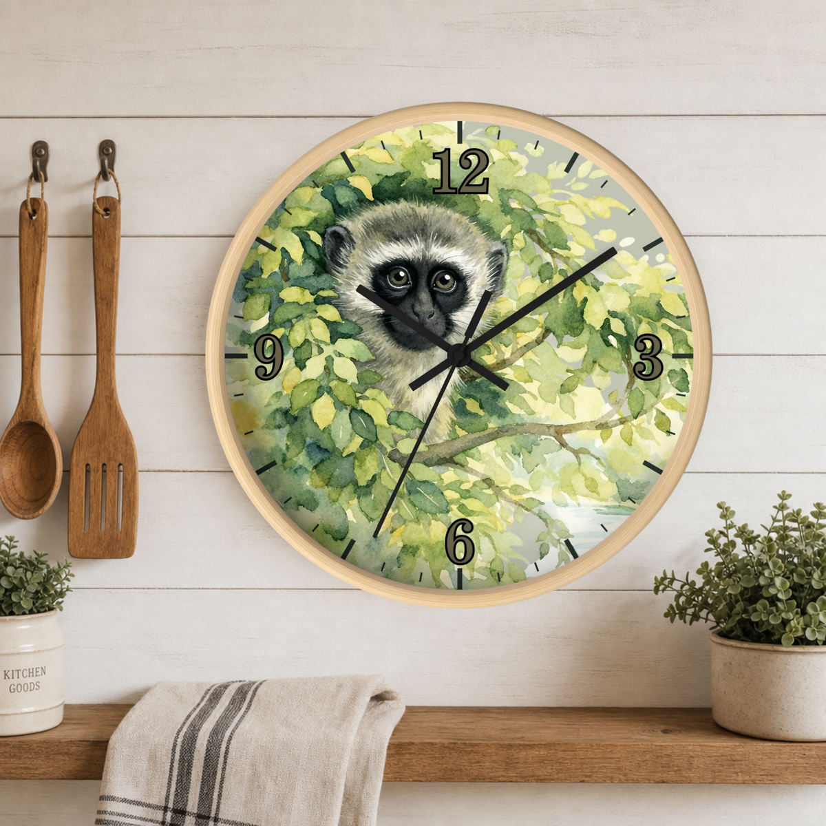 Round wall clock with a light wood frame featuring a painted vervet monkey among green leaves, mounted on a white wood plank kitchen wall above a shelf with utensils, plants, and a towel.