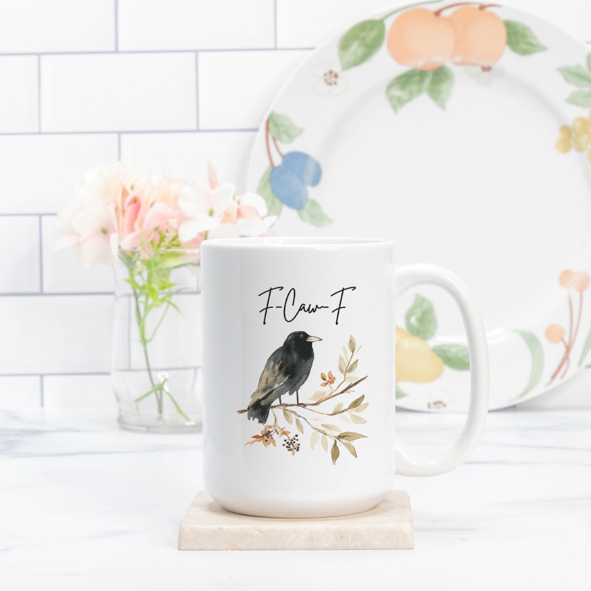 White mug with bird design and 'F.Caw.F' text on a marble coaster, with a floral-patterned plate in the background.