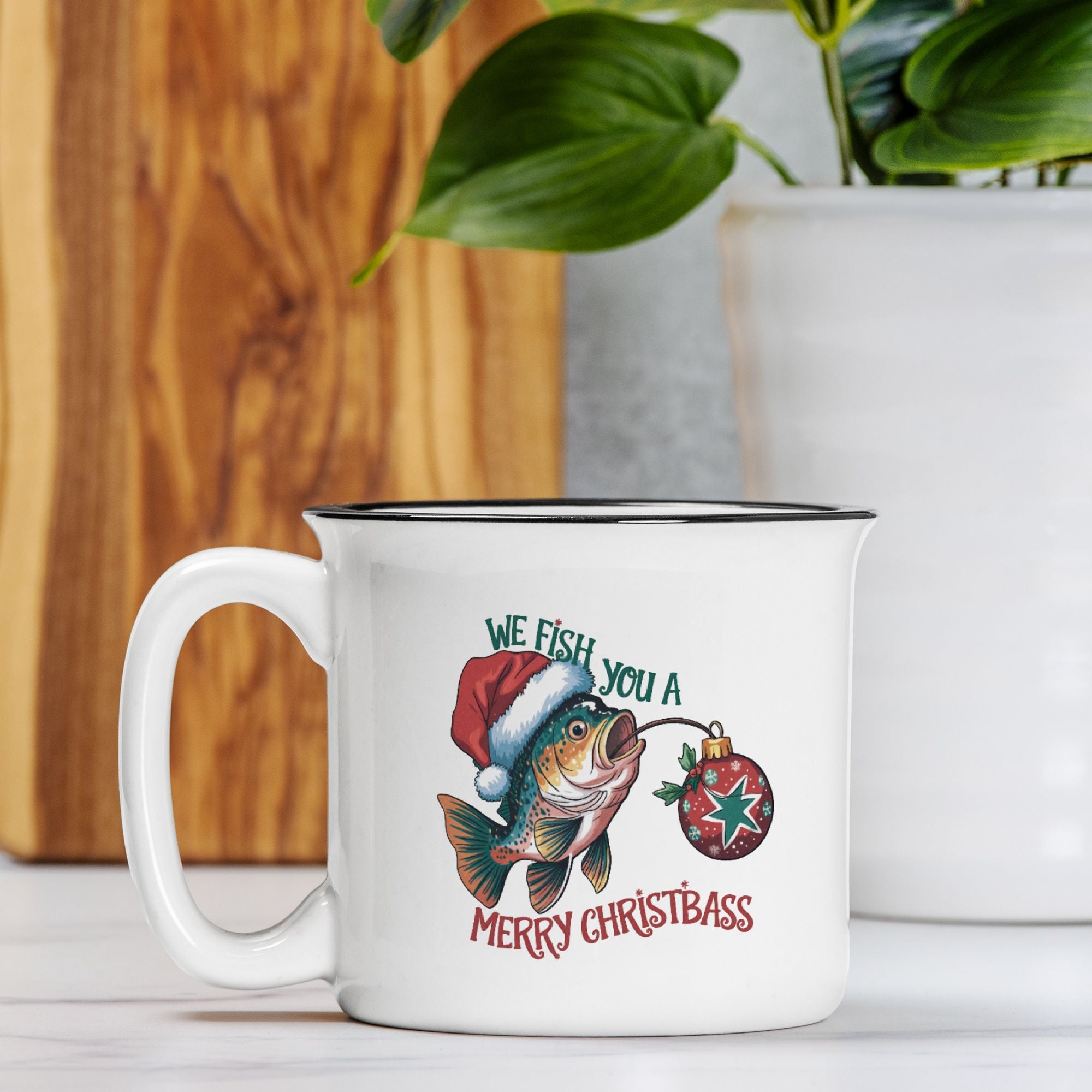 White mug with Christmas-themed design featuring a fish wearing a Santa hat and ornament, on a wooden surface with a plant in the background.