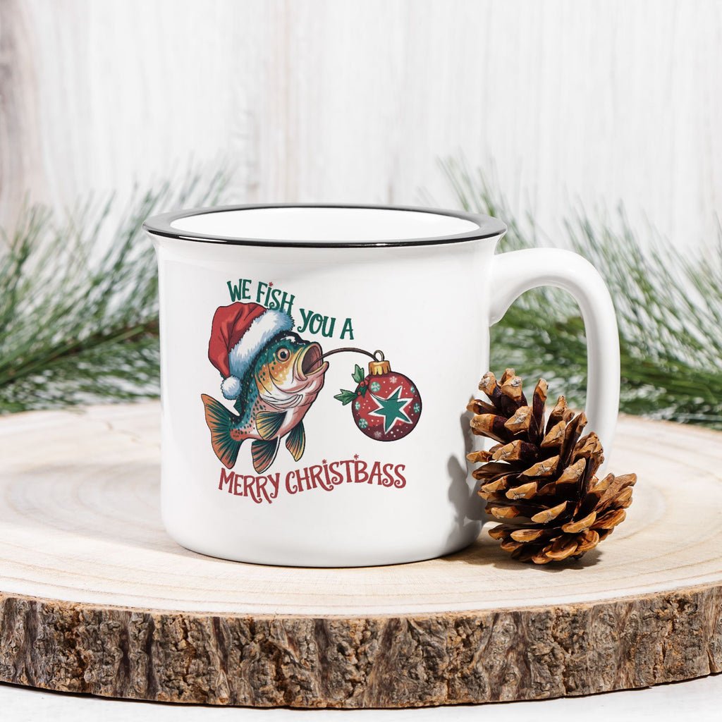 White mug with Christmas-themed design featuring a fish and Santa hat, placed on a wooden surface with a pine cone and Christmas decorations.