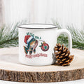 White mug with Christmas-themed design featuring a fish and Santa hat, placed on a wooden surface with a pine cone and Christmas decorations.