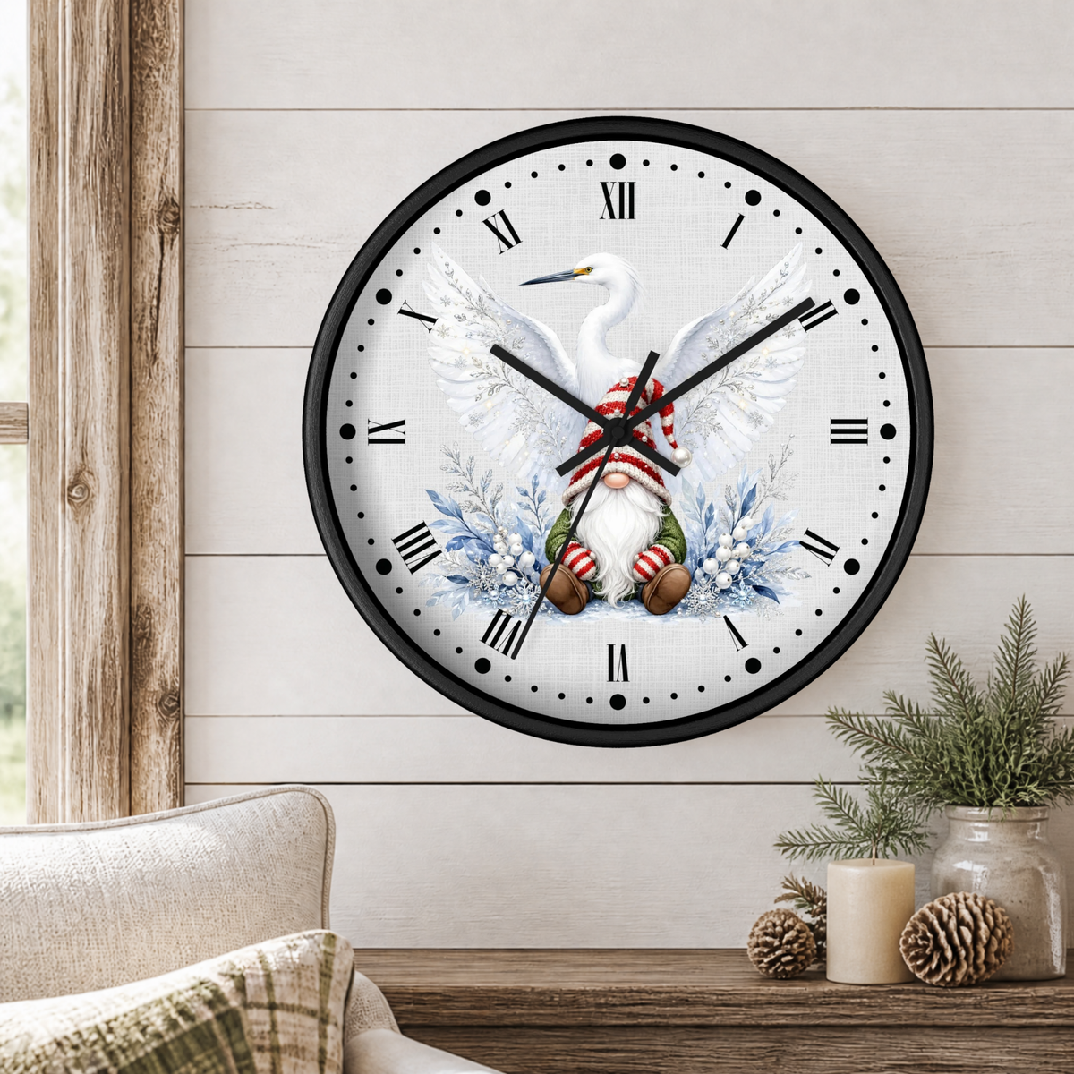 A round wall clock with a black frame and a white face featuring a white bird with wings spread, blue floral accents, and a gnome in a red-and-white striped Santa hat. The clock is mounted on a light wood shiplap wall above a wooden table with a candle, pinecones, and greenery in a styled interior setting.